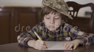 可爱有趣的小男孩穿着格子衬衫，戴着他父亲的军帽，坐在桌子旁的家里写着<strong>一封信</strong>